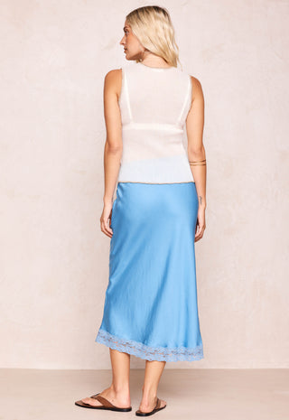 Back view of a woman with blonde beachy hair standing against a beige wall wearing a blue midi skirt with lace trim and a white tank top with brown leather sandals.