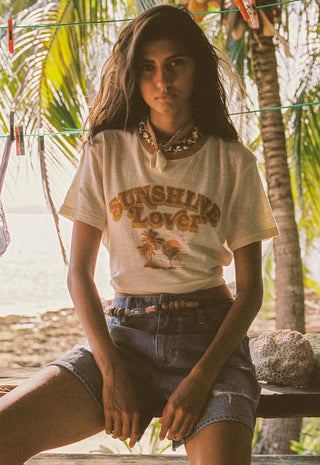 A woman sitting outdoors on a wooden bench wearing a cream graphic t-shirt with 'SUNSHINE LOVER' printed on the front, paired with denim shorts and layered necklaces, with palm trees and a beach in the background.