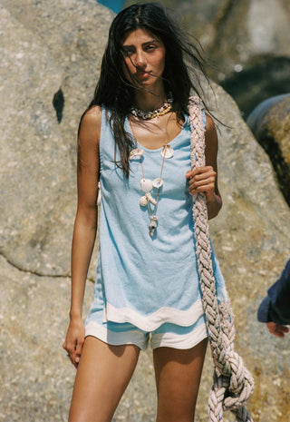 A woman standing outdoors against large rocks, wearing a light blue sleeveless top with white trim and white shorts, holding a thick braided rope. She is accessorized with layered shell necklaces.