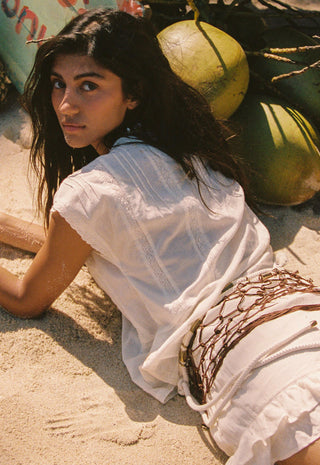 A woman lying on sandy ground wearing a white embroidered sleeveless top and skirt with a brown net belt, with coconuts in the background.