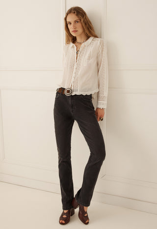 a-woman-modeling-a-white-long-sleeve-lace-blouse-tucked-into-dark-charcoal-jeans-with-a-brown-belt-and-brown-sandals-standing-against-a-white-paneled-wall