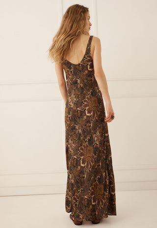 A woman wearing a sleeveless, floor-length brown maxi dress with a floral pattern, shown from the back against a white paneled wall.