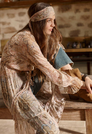 a-woman-in-a-bohemian-style-outfit-with-a-paisley-print-long-sleeve-top-and-matching-headband-leaning-on-a-rustic-wooden-table-in-a-warm-indoor-setting