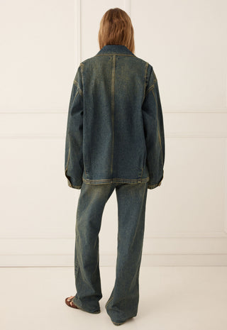 a-model-viewed-from-the-back-wearing-an-oversized-dark-blue-denim-jacket-and-matching-wide-leg-denim-jeans-standing-against-a-white-paneled-wall