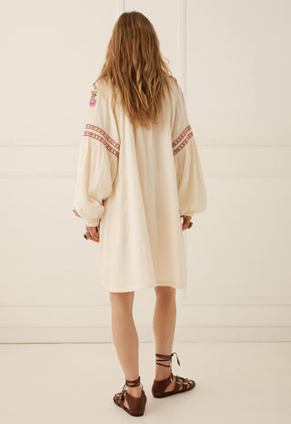 a-woman-viewed-from-behind-wearing-a-cream-colored-bohemian-long-sleeve-tunic-dress-with-red-and-orange-geometric-embroidery-on-the-sleeves-paired-with-brown-strappy-sandals-set-against-a-white-paneled-wall