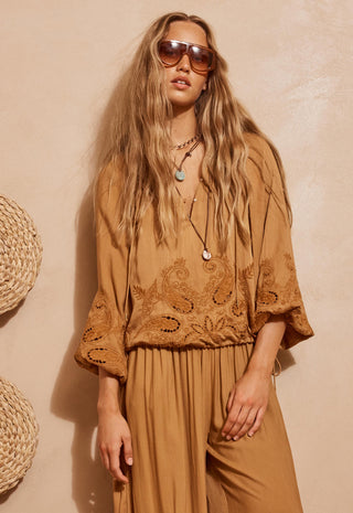A woman modeling a bohemian-style tan jumpsuit with intricate embroidery and lace detailing on the bodice and wide sleeves, styled with layered necklaces and sunglasses against a textured beige wall.
