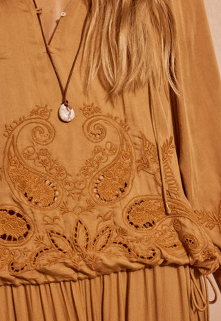 Close-up of a tan embroidered dress with paisley and floral cutout embroidery, featuring a brown cord necklace with a round shell pendant worn by a model.