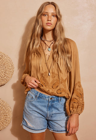 A woman modeling a tan bohemian long-sleeve blouse with intricate floral embroidery and eyelet cutouts, paired with light-wash denim shorts, set against a textured beige wall with woven baskets.
