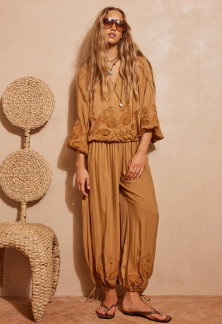 A woman models a matching tan embroidered two-piece set consisting of a loose long-sleeve top and high-waisted harem pants, styled with sunglasses, layered necklaces, and flip-flops against a textured beige wall next to a woven chair.