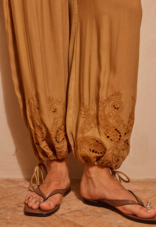 Close-up of a person wearing loose tan harem-style pants with embroidered paisley detailing near the ankles, paired with brown thong sandals on a warm beige stone floor.