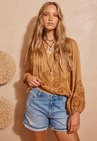 A woman modeling a tan bohemian-style long-sleeve blouse with intricate embroidery and sheer puff sleeves, paired with light-wash denim shorts, set against a warm beige textured wall with woven decorative elements.