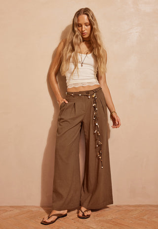 A woman stands against a textured beige wall wearing wide-leg brown pants with a decorative hanging chain detail at the side, a white cropped tank top, and black sandals.