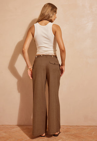 A woman shown from the back wearing wide-leg brown trousers and a white ribbed tank top, standing against a textured beige wall.