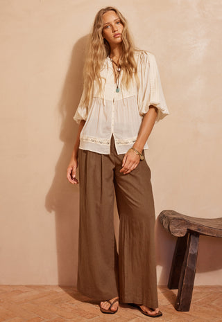 A woman stands in a warm-toned room wearing a loose white blouse with lace trim and wide-leg brown pants, paired with sandals. A rustic stool is visible against a textured beige wall.