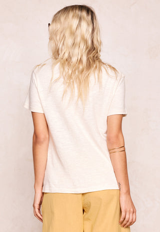 A woman shown from behind wearing a short-sleeve cream-colored t-shirt and mustard yellow pants, standing against a textured light beige wall.