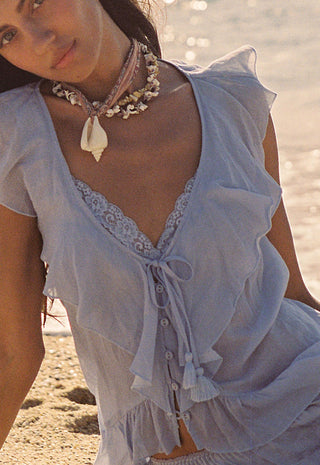A woman on a sandy beach wearing a light blue ruffled cover-up top with lace trim and a shell necklace.
