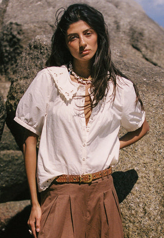 A woman wearing a white linen shirt with lace trim, brown pants, and a studded belt, accessorized with a chunky gold necklace.