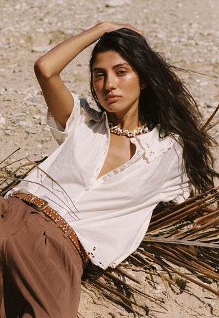 A woman reclining outdoors on a sandy ground wearing a white linen shirt with lace trim, brown pants, and a studded belt, accessorized with a chunky gold necklace.