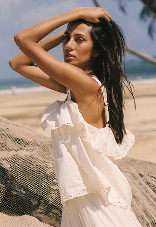 A woman on a sandy beach wearing a white ruffled sleeveless dress, with her arms raised to her head, with the ocean and sky blurred in the background.