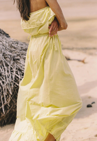 A woman wearing a flowing pale yellow maxi dress standing on a sandy beach next to a thatched structure.