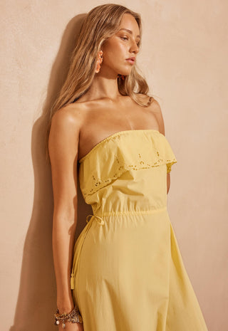 A woman modeling a strapless yellow maxi dress with ruffle details, standing against a textured beige wall in a warm, rustic setting.