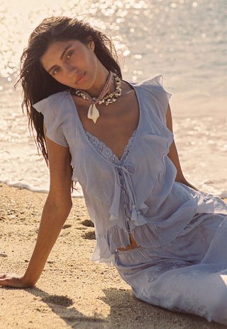 A woman sits on a sandy beach wearing a light blue sheer ruffled two-piece outfit with lace trim and a matching necklace.