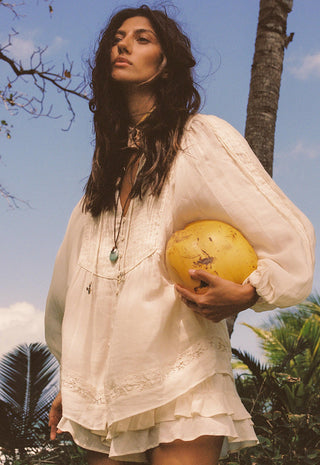 A woman outdoors wearing a loose white bohemian-style blouse and holding a yellow coconut under her arm, with palm trees and blue sky in the background.