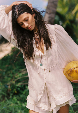 A woman outdoors wearing a loose, light cream boho-style blouse with long sleeves and a ruffled hem, holding a yellow coconut. She is surrounded by green foliage and a palm tree in a tropical setting.