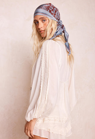 A woman wearing a white long-sleeve lace-trim blouse and a blue paisley bandana headscarf, photographed against a textured beige wall.