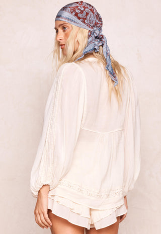 A woman shown from the back wearing a cream-colored long-sleeve blouse with lace trim and matching ruffled shorts, paired with a blue and red paisley bandana headscarf, against a textured beige wall.