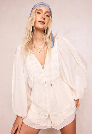 A woman modeling a cream-colored bohemian-style two-piece set consisting of a long-sleeve button-up top and matching shorts, styled with a blue patterned headband against a textured beige wall.