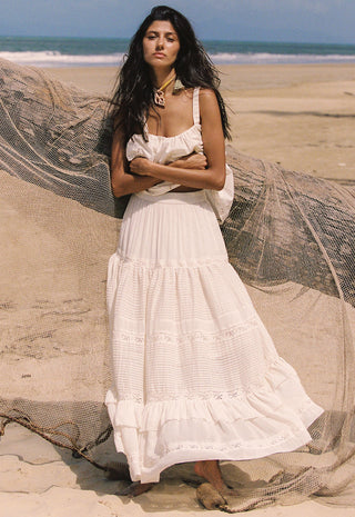 a-woman-stands-on-a-sandy-beach-wearing-a-long-white-tiered-maxi-dress-with-ruffled-trim-a-large-beige-fishing-net-lies-behind-her-with-the-ocean-and-sky-in-the-background