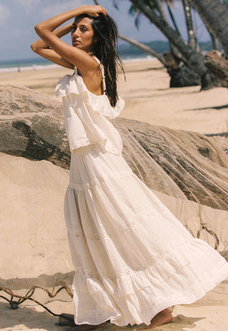 a-woman-wearing-a-long-white-tiered-boho-maxi-dress-stands-on-a-sandy-beach-next-to-a-large-driftwood-log-with-palm-trees-and-the-ocean-in-the-background