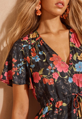A close up shot of a woman modelling a short-sleeve floral mini dress with a lace hem, standing against a textured beige wall.