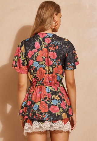 A woman shown from the back wearing a short-sleeve floral romper with a black base and vibrant pink, red, blue, and yellow flowers, featuring a lace hem.