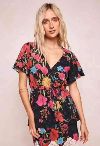 A woman modeling a black short-sleeve wrap dress with a vibrant multicolor floral print, featuring pink, red, blue, and yellow flowers with green leaves, set against a neutral beige textured background.