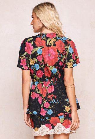 A woman shown from the back wearing a short-sleeve black dress with a vibrant red, pink, and blue floral print and a white lace hem, set against a plain beige background.