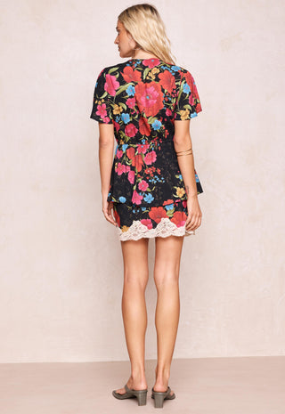 A woman viewed from the back wearing a short-sleeve black dress with a vibrant multicolor floral print and a white lace hem, paired with gray block-heel sandals against a textured beige wall.