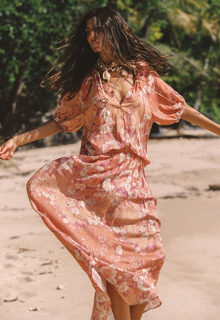 A woman wearing a flowing peach floral maxi dress with short puff sleeves and a V-neckline, captured mid-spin on a sandy beach with green foliage in the background.