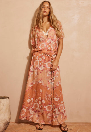 A woman modeling a peach and pink floral maxi dress with a tie waist and short flutter sleeves, standing against a textured beige wall.