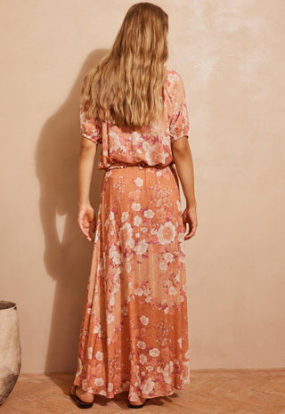A woman viewed from behind wearing a peach-orange maxi dress with a pink and white floral print, standing against a textured beige wall.