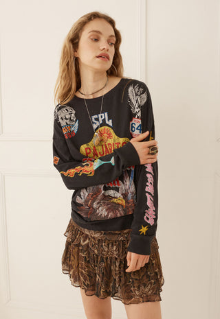 a-woman-modeling-a-black-long-sleeve-graphic-sweatshirt-with-colorful-vintage-style-prints-and-a-brown-patterned-ruffled-mini-skirt-standing-against-a-white-paneled-wall