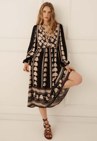 A woman modeling a black midi dress with beige floral embroidery and lace detailing, paired with brown strappy lace-up sandals, standing against a white paneled wall.