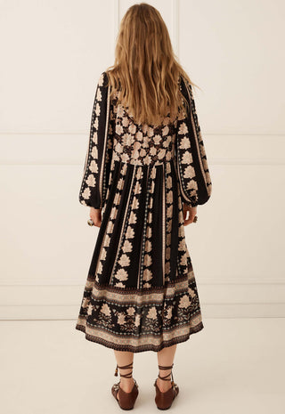 A woman viewed from behind wearing a long-sleeved midi dress with a black base and beige floral pattern, paired with brown lace-up shoes, set against a clean white paneled wall.