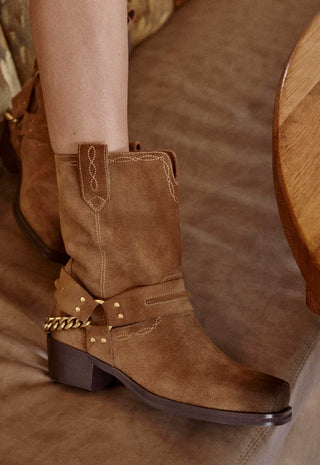 A close-up of a person's leg wearing a brown suede western-style boot with a dark brown block heel, decorative stitching, and a gold chain detail on the side, set against a brown leather couch.