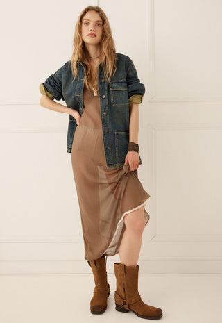 A woman models a bohemian-style outfit featuring a brown silk slip dress, a blue denim jacket with olive green rolled-up sleeves, and brown suede boots, set against a clean white paneled wall.
