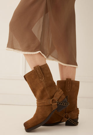 A pair of brown suede ankle boots with decorative straps and chain details, shown on a model wearing sheer brown draped pants against a light cream background.