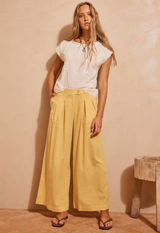 A woman stands in a studio wearing a white short-sleeve top and wide-leg yellow pants, with sandals and a necklace, against a warm beige wall.