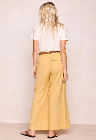 A woman shown from the back wearing wide-leg mustard yellow pants, a white t-shirt, and brown sandals against a textured beige wall.