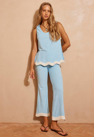 A woman modeling a light blue two-piece lounge set with white contrast trim, consisting of a sleeveless top and matching cropped pants, standing against a textured beige wall.